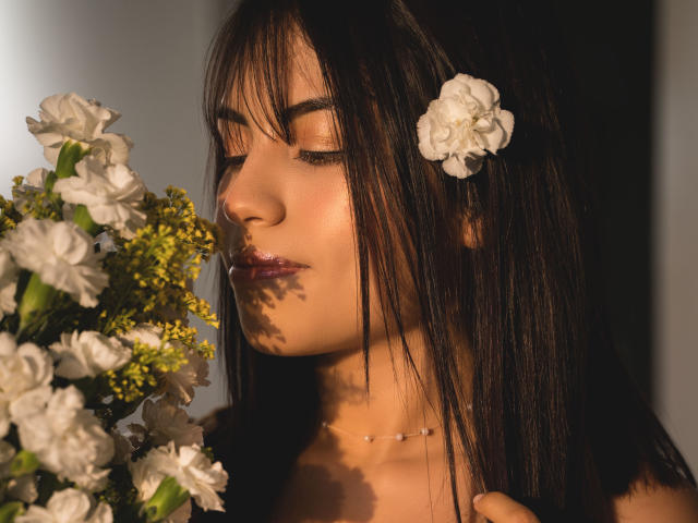 VanityValos com cabelo castanho escuro, uma flor branca no cabelo, olhos fechados, segurando flores brancas e amarelas, luz solar suave, lábios rosados brilhantes, colar delicado, pele clara, expressão suave..