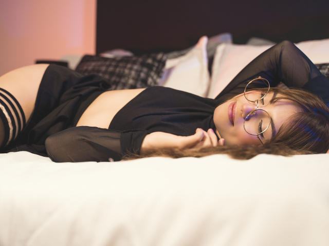 VanityValos è sdraiata su un letto, indossa biancheria intima nera con calze alte a righe, i suoi occhiali e i capelli castani sono in primo piano, una mano è dietro la testa, il sedere e i seni sono scoperti..