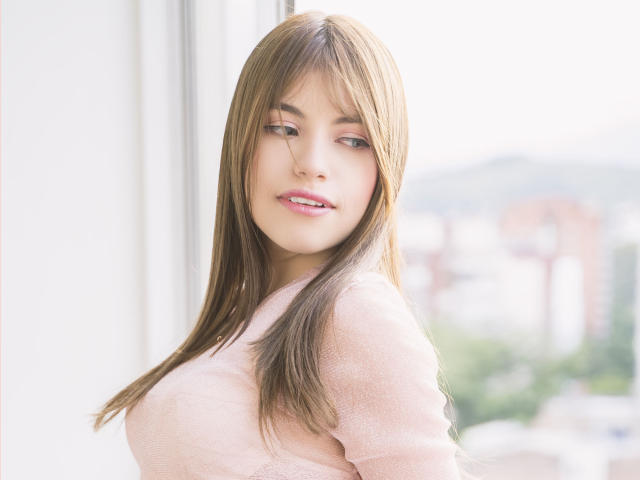 VanityValos, com cabelos castanhos longos, suéter rosa e sorriso suave, mostrando os seios, em pé perto de uma janela com uma paisagem urbana desfocada ao fundo, olhando ligeiramente para o lado..