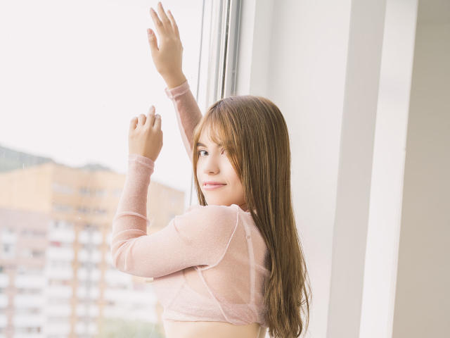 VanityValos con lunghi capelli castani, una maglietta trasparente rosa, braccia alzate, che guarda fuori dalla finestra, con un sorriso dolce, pelle chiara, seno piccolo, parete bianca, luce diurna, sfondo urbano..