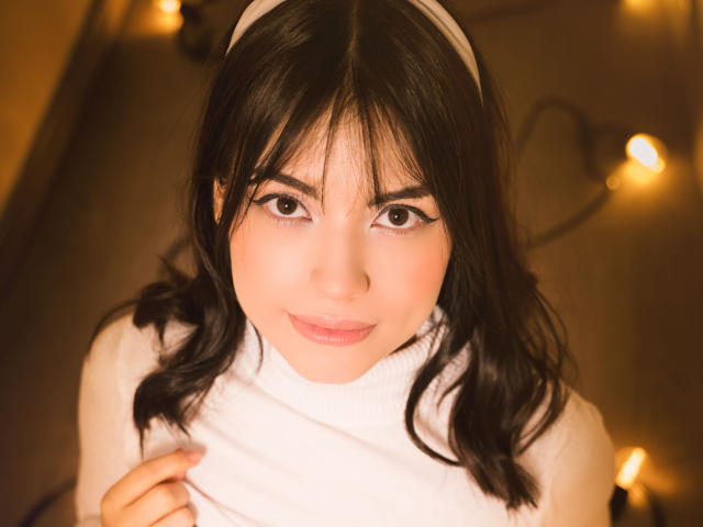 VanityValos, avec ses cheveux noirs et ondulés, son bandeau blanc et son col roulé blanc, regarde vers le haut avec ses yeux marron doux et un sourire discret, tout en tenant sa chemise près de sa poitrine..