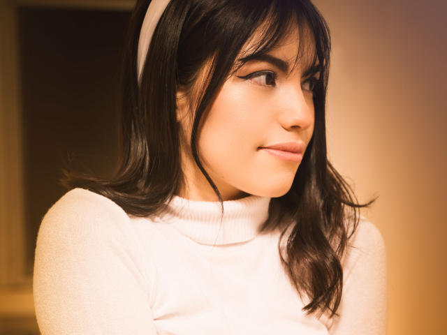 VanityValos met donkerbruin haar, een witte hoofdband en een witte turtleneck, kijkt opzij met een lichte glimlach, zacht licht op haar gezicht, warme tint, zachte uitdrukking..