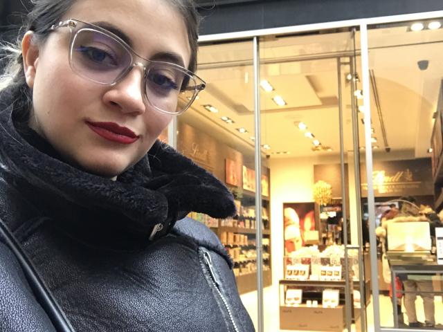 Arleen Hunter met rode lippenstift, een zwarte coltrui en een bril, een zwarte leren jas, staand voor een winkel met glazen deuren, kijkend in de camera, lachend, bruin haar in een paardenstaart..