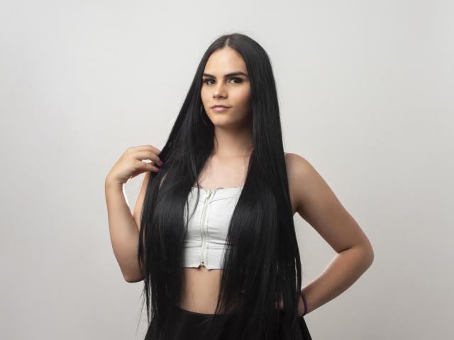 JassmineTS met lang zwart haar, draagt een witte crop top, haar hand in haar haar, met een zelfverzekerde blik, staand voor een effen achtergrond. De borsten zijn bedekt door de crop top..