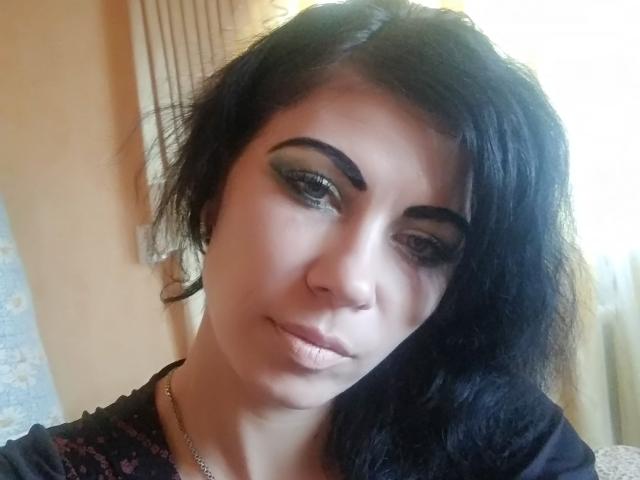 JuliaIce, avec ses cheveux noirs, son fard à paupières vert et ses lèvres pulpeuses, porte un haut noir avec des motifs floraux roses, et regarde légèrement sur le côté avec un sourire discret..