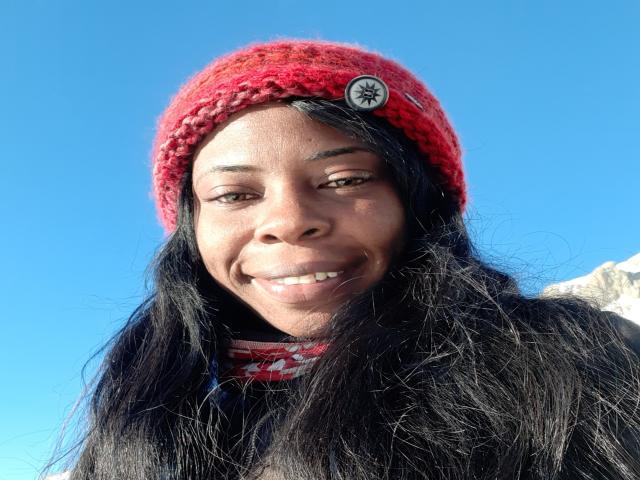 LencyHot with dark skin, long black hair, red knitted hat with button, smiling, wearing black top, against clear blue sky. Boobs not visible, focus on face and hat.