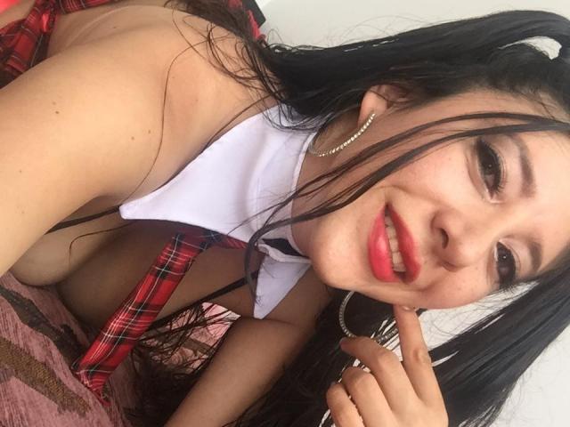 HotPamelaSex with black hair, red plaid schoolgirl outfit, white shirt, and red lipstick, lying on pink bed, touching chin, smiling, wearing hoop earrings, showing her tits.