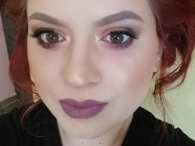 AnaisGrosSeinss ha la pelle chiara, i capelli rossi, le sopracciglia folte, l'eyeliner scuro, l'ombretto viola, gli occhi verdi e il rossetto viola. Il suo viso è al centro dell'immagine..