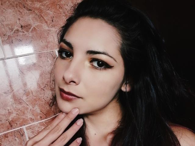 SanraX hat dunkle Augen mit auffälligem Eyeliner, volle Lippen mit einem rosa-roten Lippenstift und langes, schwarzes Haar. Ihre Hand ruht sanft auf ihrem Hals und zeigt ihre gepflegten Nägel..