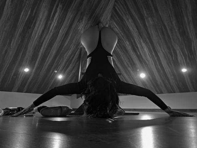 MiaBlackXO sta eseguendo la posizione dello yoga del "cane a testa in giù", con i glutei sollevati, indossando un body nero e con la testa tra le gambe, in una stanza buia con pannelli di legno..