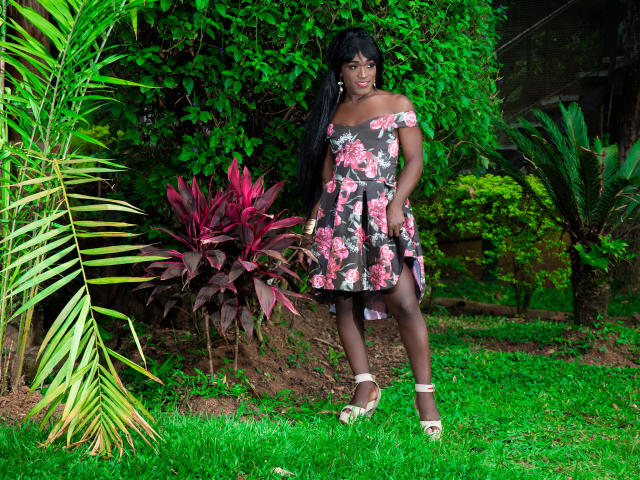 LatinWonderCockTS em um vestido floral com um ombro de fora, mostrando seus seios, pele negra, cabelo preto longo, sandálias brancas, em pé na grama verde, com plantas vermelhas e verdes..