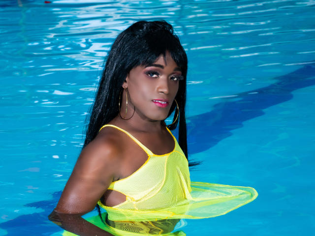 LatinWonderCockTS con pelle scura, capelli neri e rossetto rosa, che indossa una maglietta gialla in rete, in piedi in una piscina di un blu brillante, che guarda la fotocamera con un leggero sorriso..
