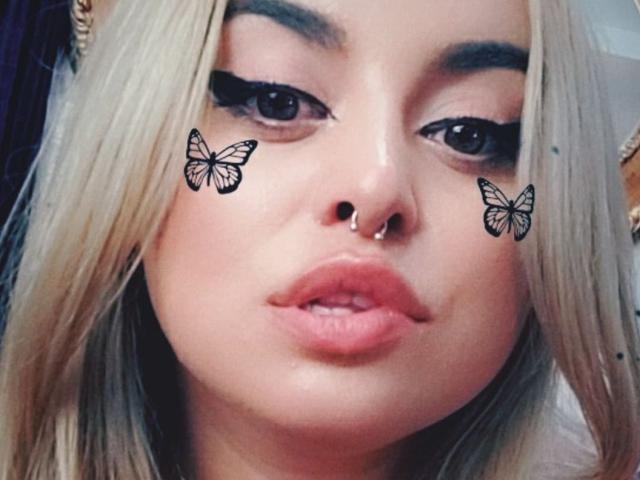 ChaudePourxToi with blonde hair, black eyeliner wings, butterfly tattoos on cheeks, nose ring, and pink lips. Her eyes are dark and she has a slightly pouty expression.