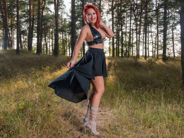 IvyFox with red hair, wearing a black leather bra, high-waisted skirt, and silver strappy heels, standing in a forest, holding her skirt, showing her toned ass.