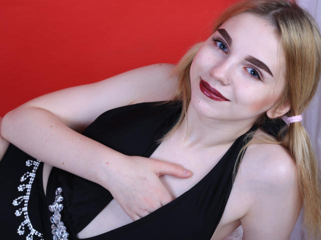 ScarletAmi avec des couettes blondes, du rouge à lèvres rouge foncé et une robe noire, pointant ses seins nus avec sa main droite, souriante, avec un anneau au nez, et un fond rouge..