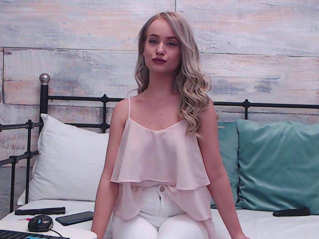 SophieXDee with blonde curls, wearing a pink ruffle top and white pants, sitting on a bed with teal pillows, and a black metal headboard, looking at the camera.