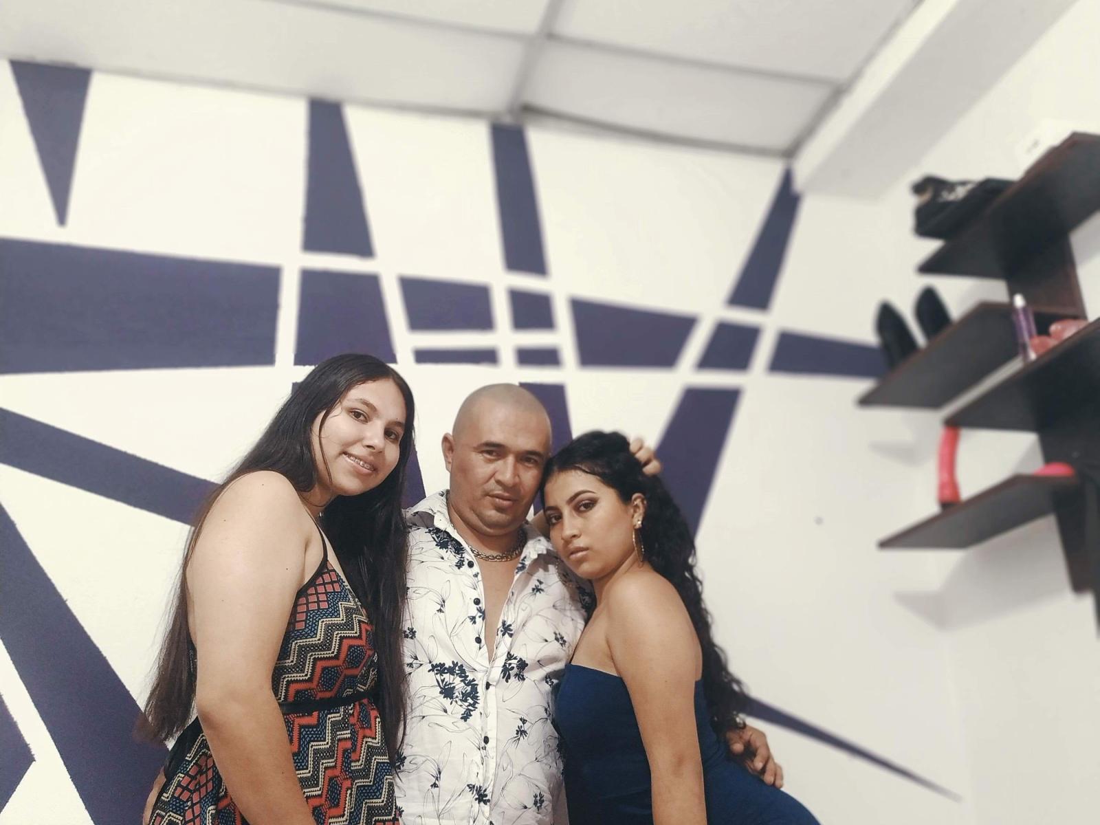Kratos, Xiommy, Erica, com a cabeça raspada, camisa branca com estampa floral, cercadas por duas mulheres, uma vestindo um vestido com padrão em zigue-zague e a outra com um vestido azul sem alças, todas em pé, com os braços ao redor umas das outras, e expressões sérias..