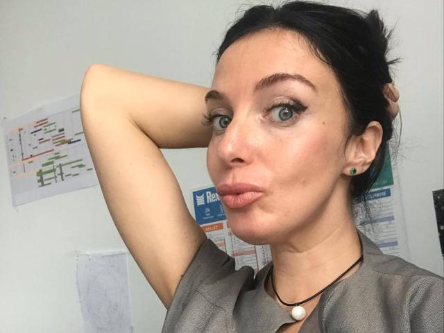 KatieFrenchie has puffy ass-free lips, green eyes, black hair in a messy bun, wearing a grey shirt, black necklace with a white bead, and green earrings.