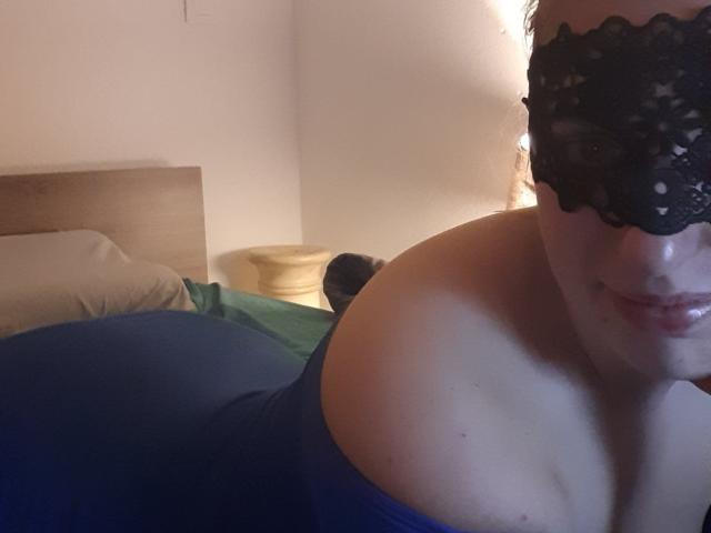 El rostro de Francaise, con un antifaz de encaje negro, una blusa azul con un hombro descubierto que muestra los hombros desnudos y la parte superior del pecho, con una ligera sonrisa, en un dormitorio tenuemente iluminado..