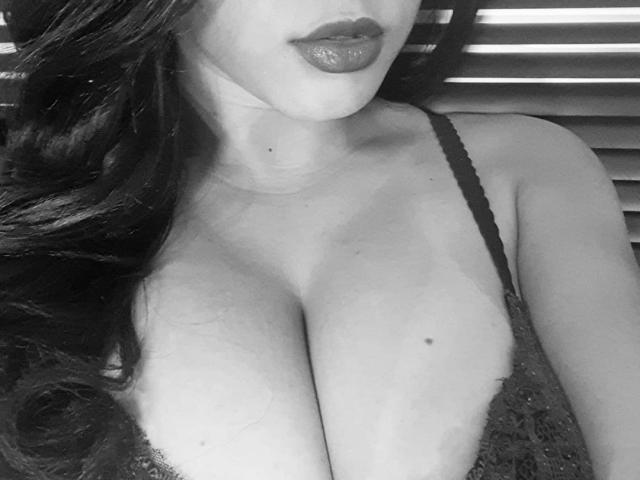 MagalySexy's large tits in a black lace bra, with long wavy dark hair and glossy lipstick, in a black and white photo. Her cleavage is prominent and she's slightly tilted.