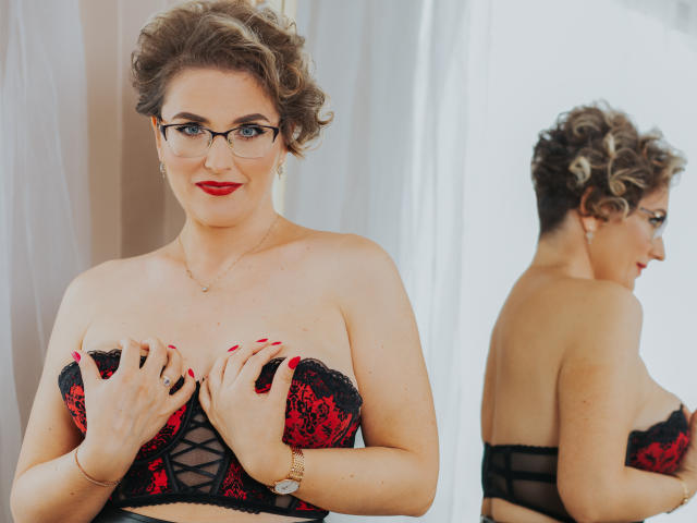 Cara Jones, con capelli castano ricci, rossetto rosso, occhiali, reggiseno di pizzo nero e rosso, mani sulle tette, riflesso nello specchio che mostra la schiena. Sta sorridendo, unghie rosse, orologio d'oro..