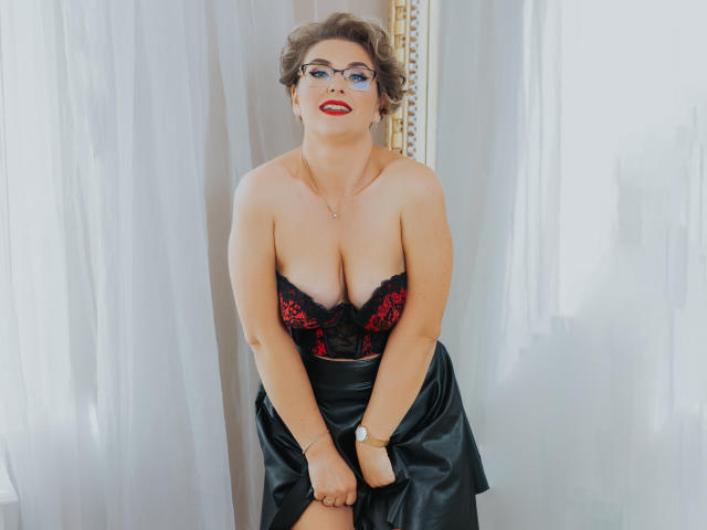 CaroJones with curly brown hair, glasses, red lipstick, black lace bra showing big tits, and black satin skirt, sitting with hands on knees, wearing a gold watch.