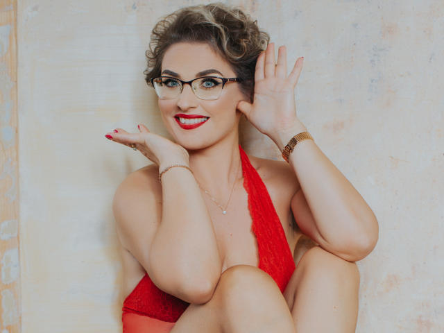 CaroJones with curly brown hair, glasses, red lipstick, and red lace halter top, smiling, waving with hands on cheeks, wearing a gold bracelet, sitting with knees up.