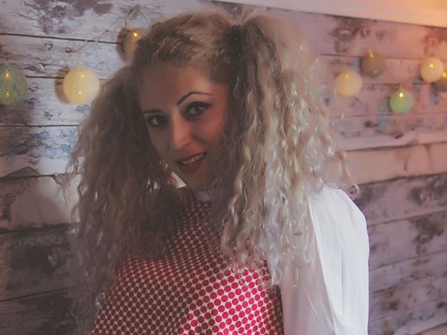 SassyKate with curly blonde pigtails, smiling, wearing a white shirt and red checkered dress, has a cute face and small tits. Warm, soft lighting. Cozy, rustic setting.