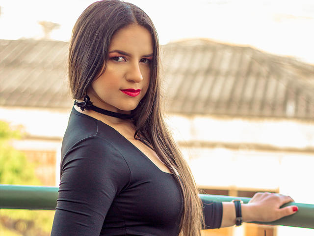 AbbyShiine with long brown hair, black choker, and red lipstick, wearing a tight black top, showing her small tits, standing by a green railing, smiling confidently.