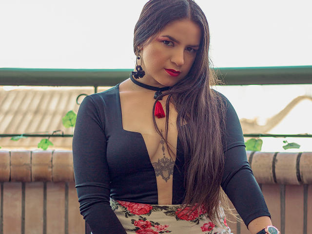 AbbyShiine has long brown hair, red lipstick, black choker, and deep V-neck black top showing her tits with a tattoo below. Floral skirt, earrings, and confident gaze.