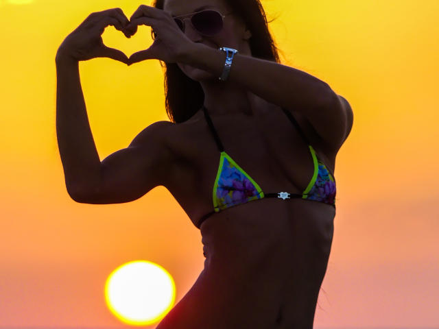 Silhouetted CassandraMichelli with toned abs, wearing a colorful triangular bikini, making a heart shape with her hands, sunset in the background, wearing sunglasses and a bracelet.