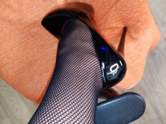 AnabelSex69s Fuß in schwarzen Fischstrumpfhosen befindet sich in ihrem Mund, wobei ein glänzender schwarzer High Heel-Schuh teilweise darin steckt. Ihre Lippen umschließen die Spitze des Absatzes..
