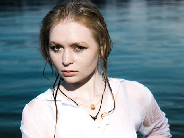 SweetAleksaX with wet, blonde hair, pale skin, and a serious expression, wearing a white, wet shirt, and a necklace with a small wooden pendant. Blue water in background.