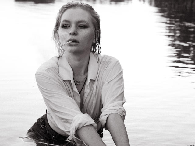 Zwart-wit foto van SweetAleksaX met nat blond haar, die een doorweekt wit shirt en donkere broek draagt, zittend in het water, een sigaret rookt, en er moe en aantrekkelijk uitziet..
