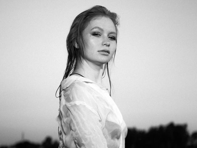 Black-and-white photo of SweetAleksaX with wet, dirty blonde hair, wearing a sheer, wet, patterned top, showing small, perky tits, and a confident, slightly annoyed expression.