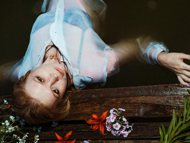SweetAleksaX com cabelo ruivo, vestindo uma camisa azul transparente, deitada na água com tábuas de madeira, flores e um colar, olhando para cima com uma expressão ligeiramente triste..