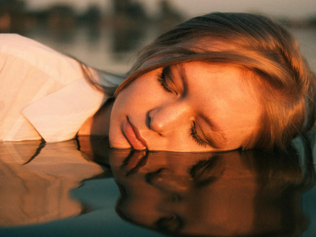 SweetAleksaX's face is partially submerged in water, eyes closed, with her reflection clearly visible. She's wearing a white shirt, and her blonde hair is wet. Golden sunlight.