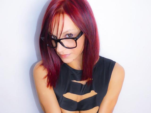 CassandraMichelli has red hair, black glasses, and a black cut-out top, showing her small tits. She's smiling slightly, with a white background.
