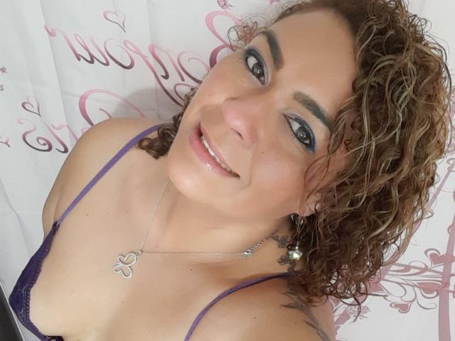 SolmaryHot with curly brown hair, purple bra, and silver necklace, smiling at the camera, showing small tits and a tattoo on her right shoulder. She has blue eyes and eyeshadow.