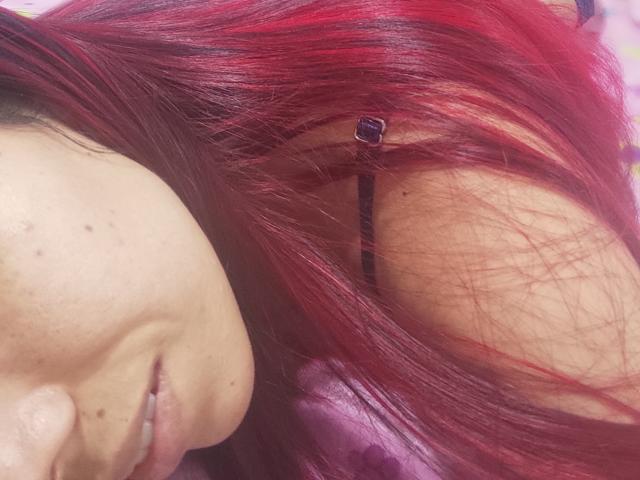 Le visage de VenussHot, avec ses cheveux roux et noirs, présente une tache de rousseur sur sa joue et une petite épingle à cheveux noire dans ses cheveux. Ses lèvres sont légèrement entrouvertes..