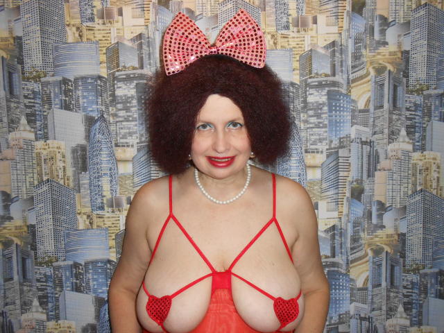 AirMagdalene con i capelli rossi ricci, un fiocco rosso a pois, una collana di perle, lingerie rossa con spalline sottili, che mostra il suo seno generoso con dei dischetti rossi a forma di cuore, sorridendo alla fotocamera..