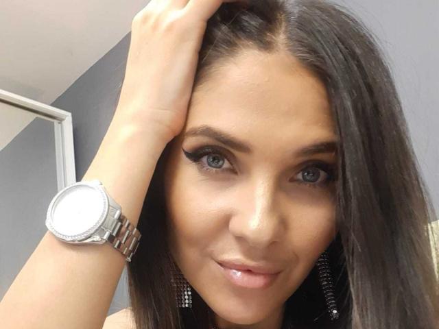 LovelyKinsley with dark hair, hand in hair, wearing a silver watch, eyeliner, and shiny pink lips, blue eyes, and small nose, smiling slightly. Boobs not visible.