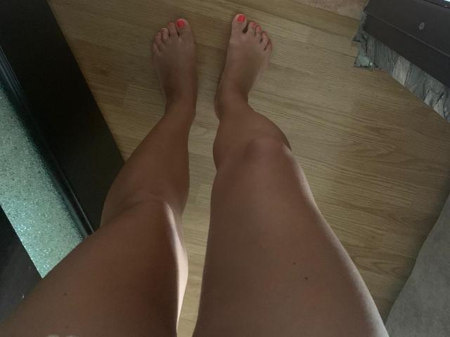 Le gambe e i piedi di TanyaMonroe sono scoperti, con le unghie dei piedi rosa, e sono in piedi su un pavimento di legno. La sua pelle è abbronzata e l'immagine è una ripresa dal basso, incentrata sui piedi..