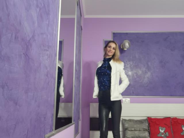 PlayfulErica with blonde hair, white jacket, blue sparkly top, black leather pants, standing in purple room, smiling at mirror, reflection showing her tits and ass.
