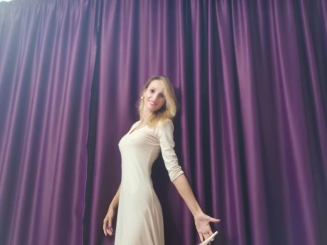 PlayfulErica stands in a purple curtain, wearing a tight, beige dress, her blonde hair down, holding white heels in one hand, smiling, with a slightly tilted, confident pose.