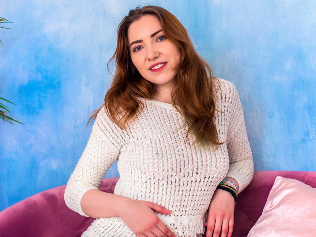 Decadance with wavy brown hair, blue eyes, and a smile, wearing a white knit sweater, sitting on a pink couch, right hand on her belly, left wrist with bracelets.