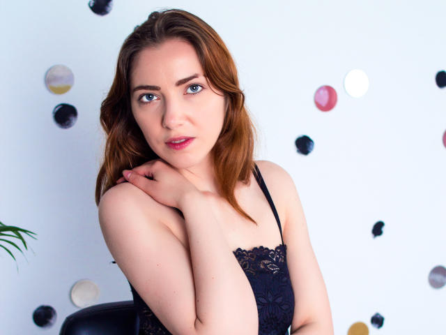 Decadance has light skin, brown hair, blue eyes, and small tits in a black lace top, looking at the camera with a slight smile, hand on her neck.