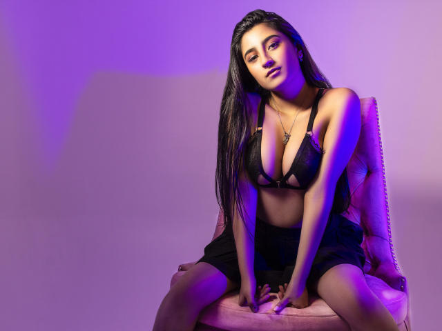 AngelaRos, con cabello negro largo, vestida con un sostén y falda negros, sentada en una silla rosa, con luces moradas y azules sobre ella, mirando ligeramente hacia un lado con una expresión relajada..