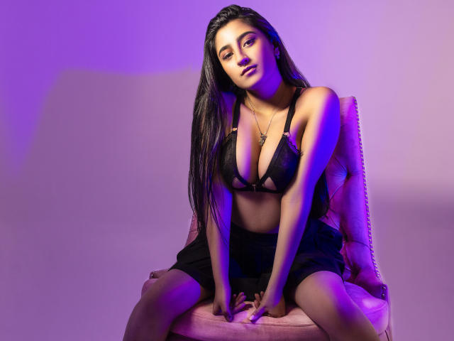 AngelaRos, com longos cabelos pretos, iluminada em tons de roxo, sentada em uma cadeira rosa, vestindo um sutiã e shorts pretos, exibindo seios grandes, nariz preto e uma expressão confiante e sexy..