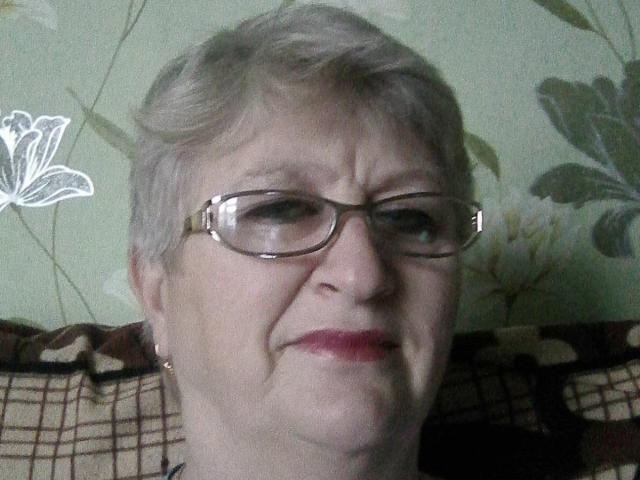 LolkaAlyss with short gray hair, glasses, and red lipstick, has a slightly annoyed or tired expression, sitting on a plaid-covered chair.